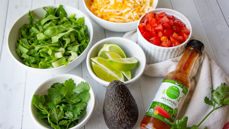 bowls of lettuces, cheese, tomatoes, lime wedges, and cilantro by an avocado and a bottle of hot sauce