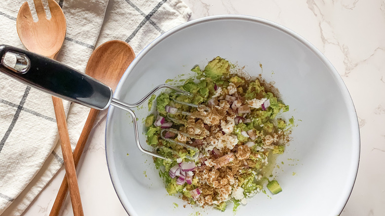 white bowl of mashed avocado filled with red onion, lime juice, ground cumin, salt, and pepper near wooden spoons on a counter