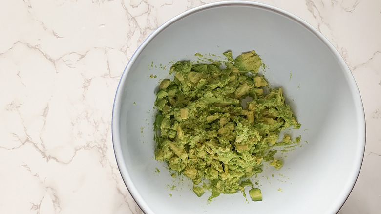 white bowl of mashed guacamole in a white bowl near a potato masher and wooden spoons on a counter