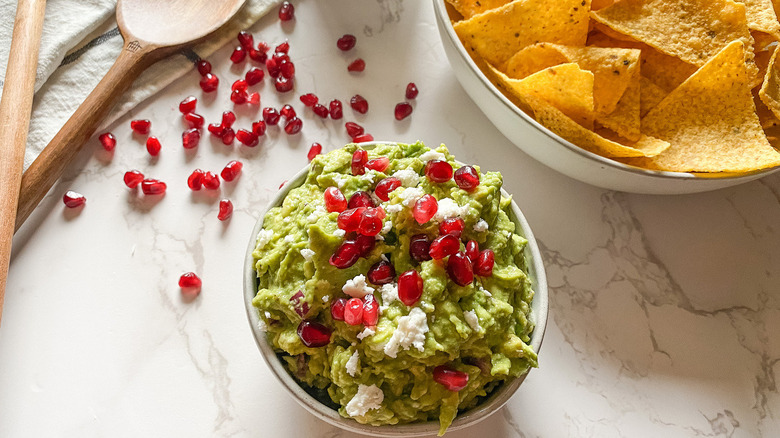 bowl of guacamole topped with pomegranate seeds near a bowl of tortilla chips and wooden spoons on a counter