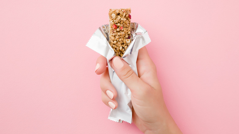 Young woman hand holding cereal bar on pastel pink table. Opened white pack.