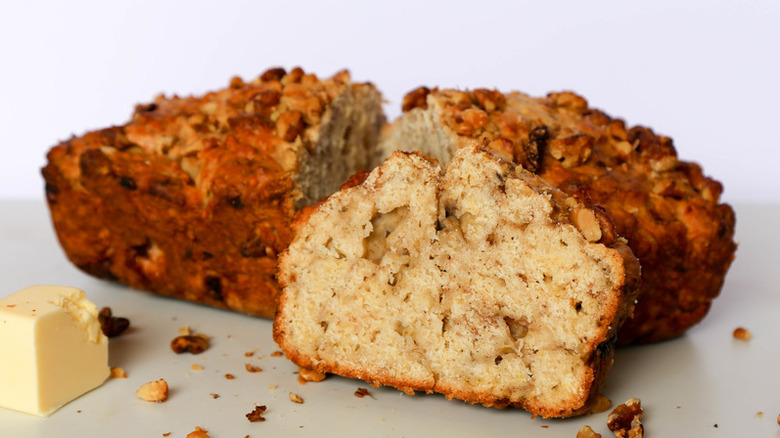 Slice of banana bread with walnuts, with loaf behind. 