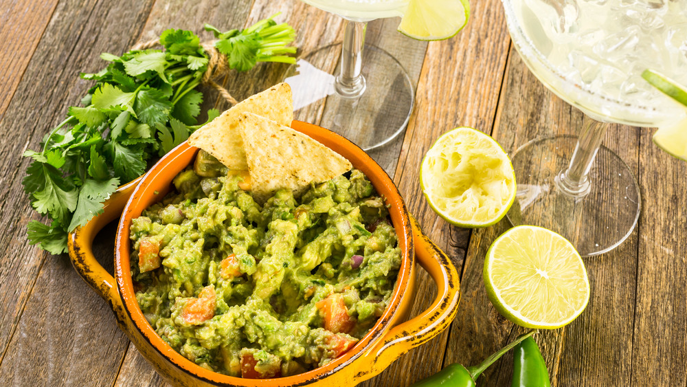 Bowl of guacamole with a couple tortilla chips, two margaritas, a lime cut in halves, cilantro, and peppers on wooden surface