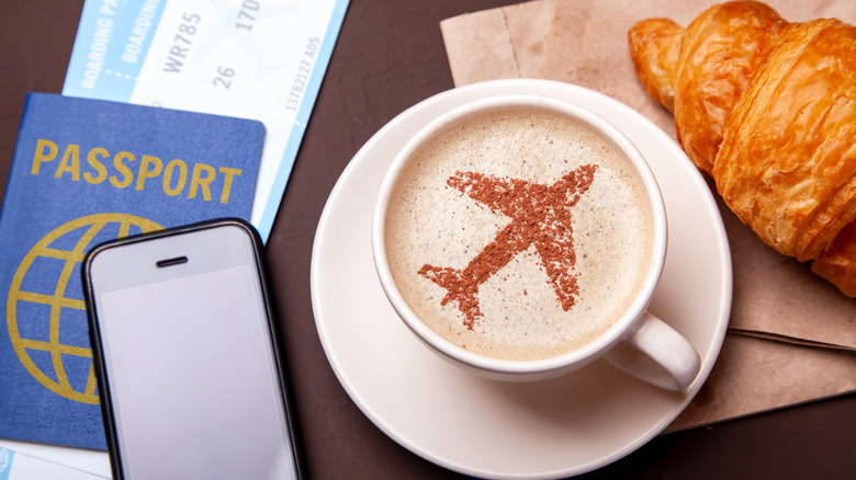 A cup of coffee next to a passport, phone, and a croissant