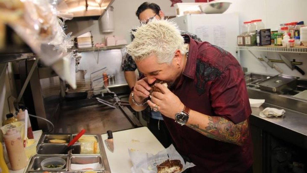 Fieri taking a large bite out of a sandwich in a restaurant kitchen