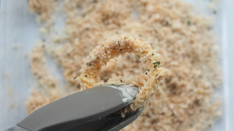 A coated onion ring held by tongs.