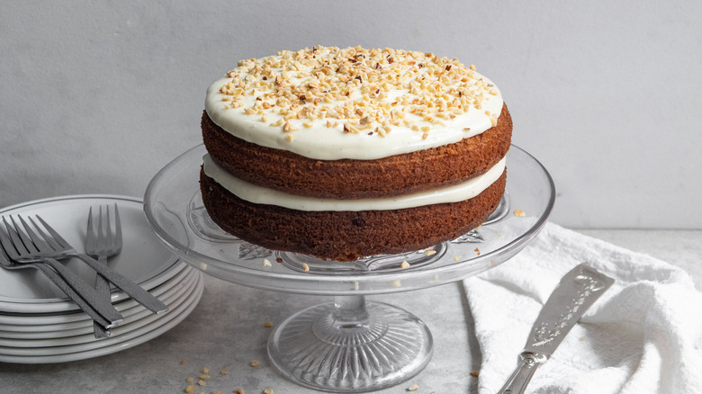 two-layer spice cake with cream cheese frosting and chopped hazelnuts on a glass cake stand near dessert plates and forks