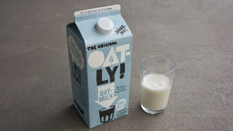 Oatly oat milk in cardboard container next to clear glass of oatmilk on gray background