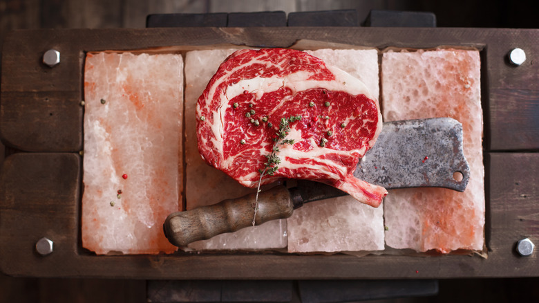 a ribeye steak sitting on salt bricks in a wooden board with a cleaver and herbs