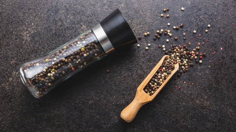 pepper mill filled with peppercorns beside fresh peppercorns on a slate background