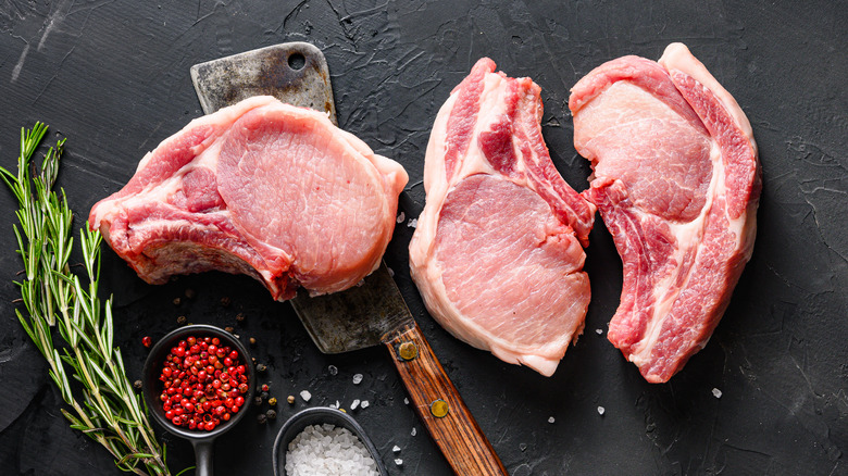 Raw pork chops beside a cleaver, fresh herbs and seasonings on a black background