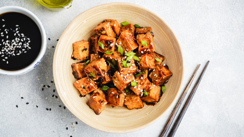 Saucy cooked tofu on tan with green onions and sesame seeds on tan plate