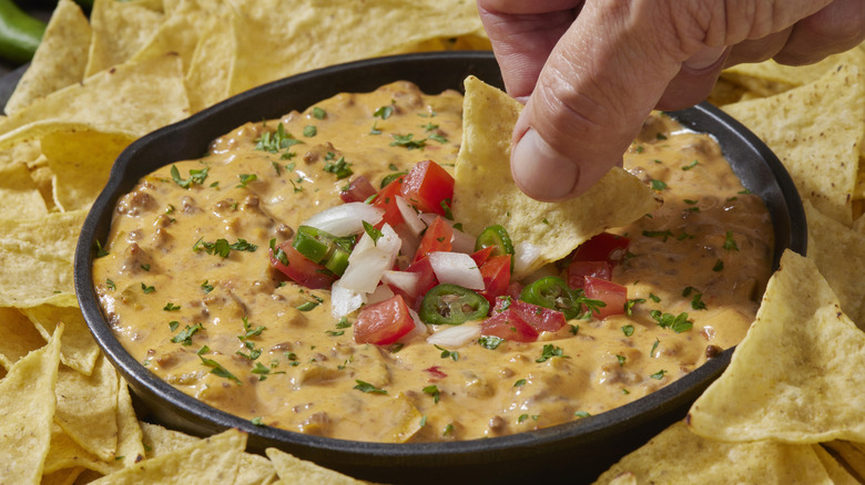 hand dipping tortilla chips into chunky nacho cheese sauce