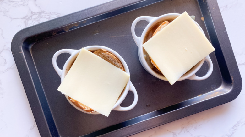 Cheese slices on French bread on top of onion soup in bowls, on a baking sheet