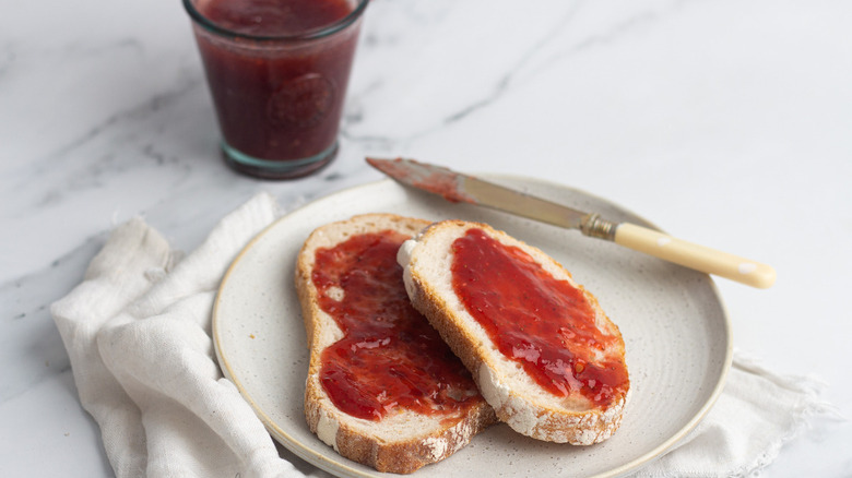 instant pot homemade strawberry jam in a glass jar and spread across two pieces of toast on a white plate near a knife