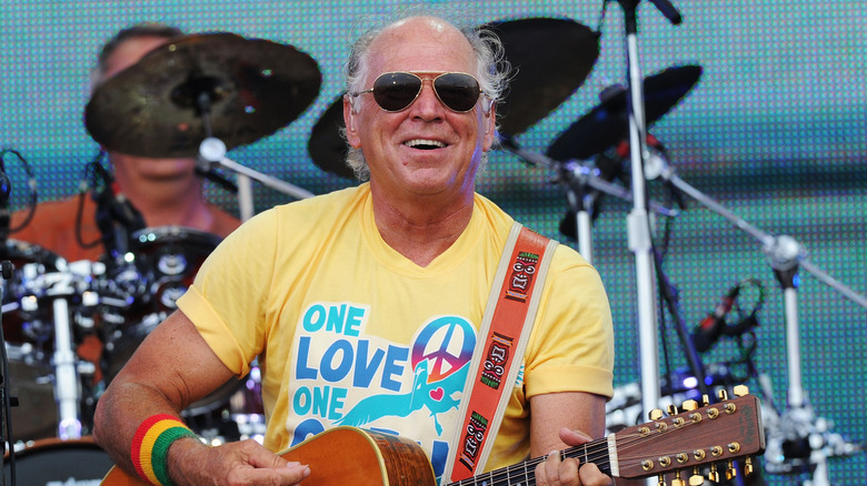 Jimmy Buffett in sunglasses, smiling and playing guitar