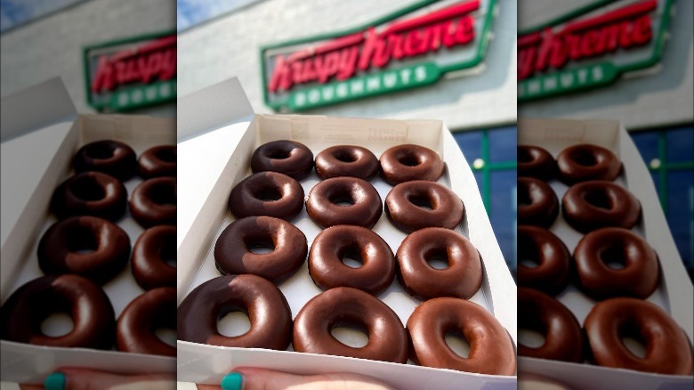 Person holding a box of chocolate glazed doughnuts in front of a Krispy Kreme