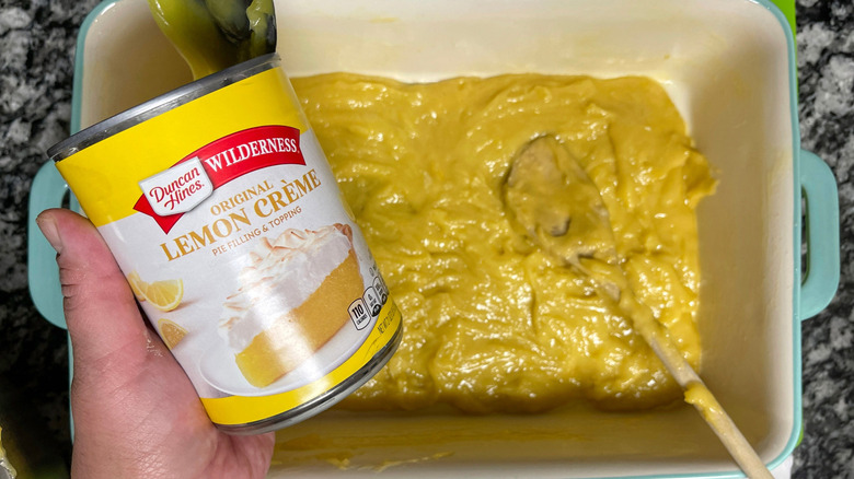 Hand holding can of lemon pie filling over batter in baking dish