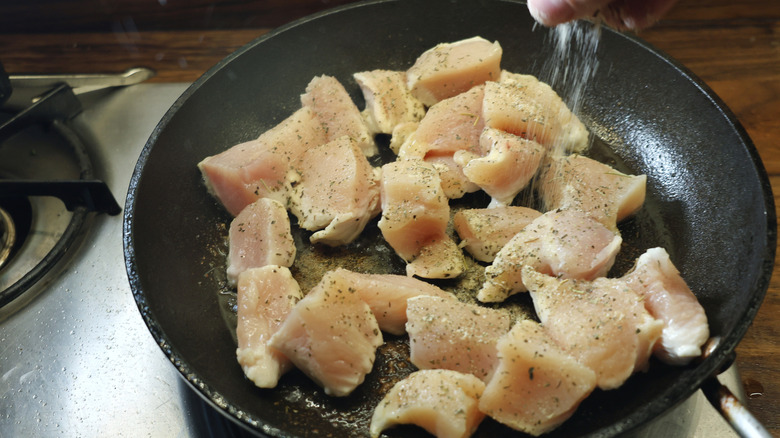 hand sprinkling seasoning over chicken breast cubes in skillet