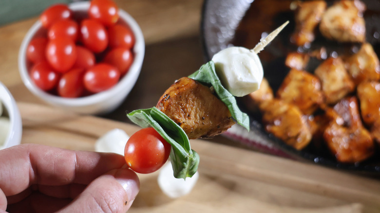 hand holding Margherita chicken skewer in front of bowl of tomatoes and skillet of balsamic-glazed chicken