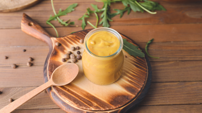 Jar of honey mustard on wooden plate with wooden spoon, on wooden table with mustard seeds and arugula leaves