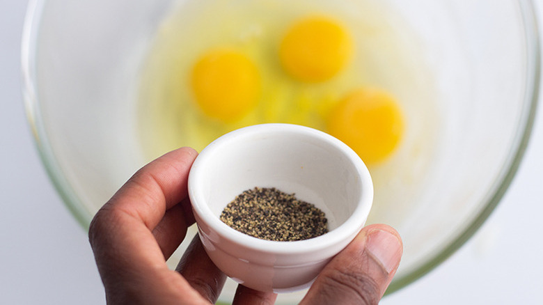 A hand holding a container of pepper over a glass mixing bowl containing egg yolks and whites