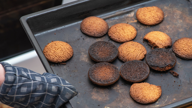 A hand with an oven mitt pulling a sheet of burnt cookies out of the oven