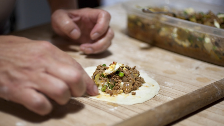 Hands adding filling to empanada dough on a wood board