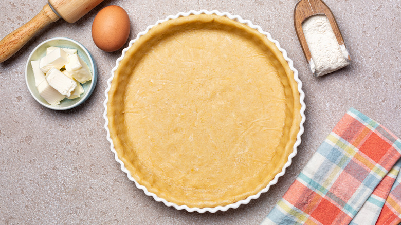 An unbaked pie crust in a pan surrounded by butter, flour, and an egg