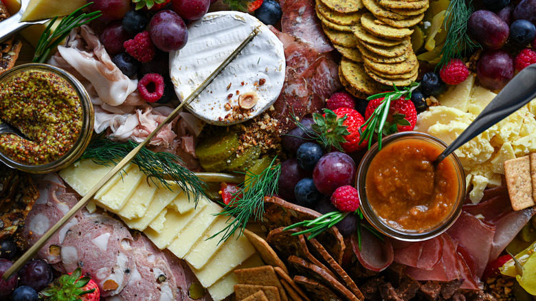 close up of charcuterie board with various meats, cheeses, fruit, and crackers plus small glass pots of grainy mustard and an orange fruit preserve