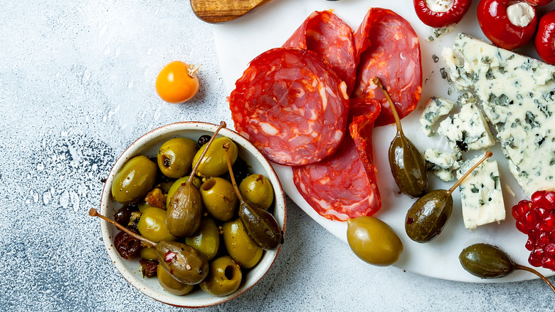 white plate with charcuterie meats and cheeses with bowls of olives and sun dried tomatoes and two glasses of white wine