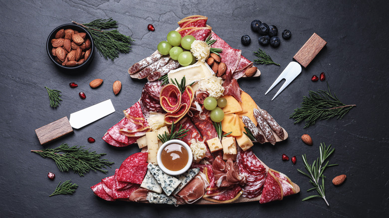 Christmas tree shaped charcuterie board with multiple types of cured meats with some cheese, fresh fruit, and nuts on dark slate background with fresh greens