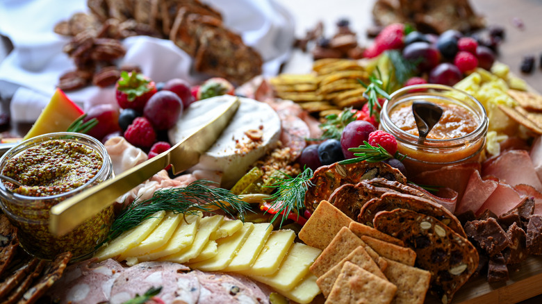 close up of charcuterie plate with meats, cheeses, crackers, wedge of brie with butter knife, and small pots of mustard and preserves with small spoons in them