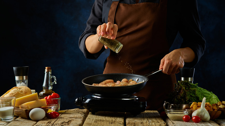 A cook pouring spices from a jar over raw poultry 