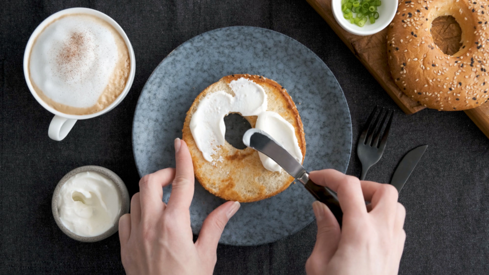 spreading cream cheese on toasted bagel with a latte drink