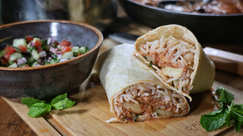 sliced butter chicken burrito on cutting board next to bowl of kachumber