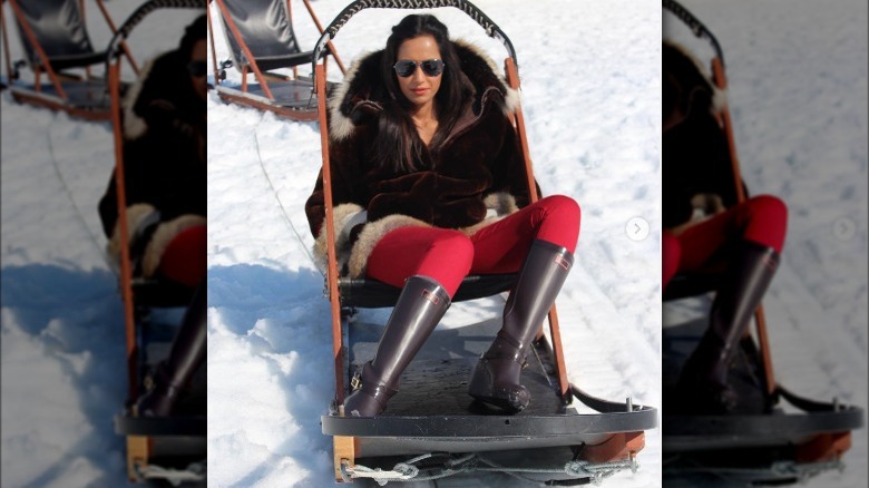 Padma Lakshmi in red pants and boots on a sleigh
