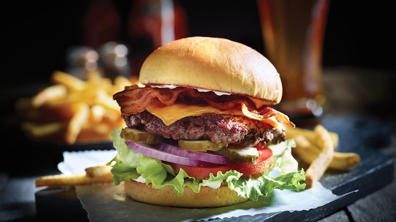 the classic bacon cheeseburger with bacon and melted cheese atop a beef patty with pickles, onion, lettuce, and a tomato served alongside seasoned fries on a gray wooden table