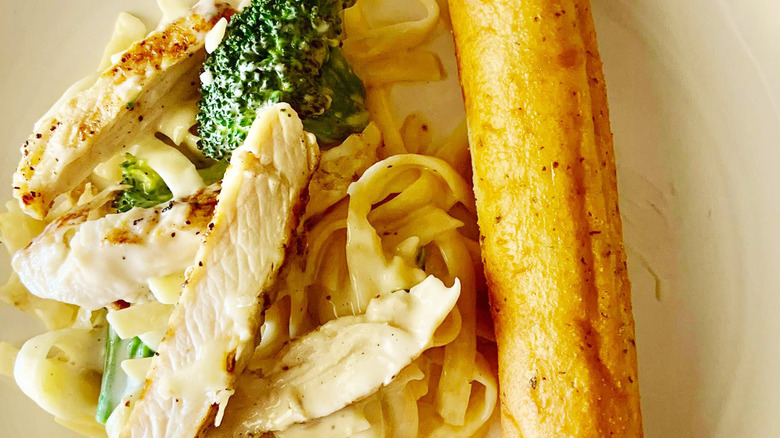 strips of grilled chicken atop a bed of fettuccine noodles with broccoli and alfredo sauce alongside a bread stick on a white plate