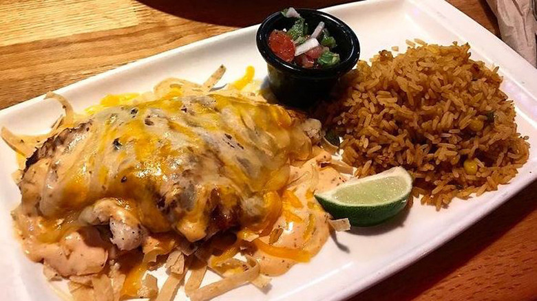 white plate of grilled chicken smothered with a blend of cheddar cheeses and a lime sauce served alongside Spanish rice and a cup of pico de gallo