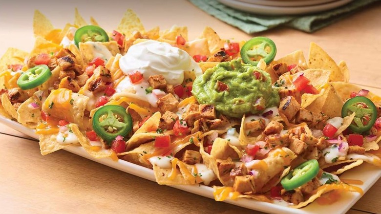 white plate full of nacho chips, jalapeno peppers, ground beef, tomatoes, onion, cheese, and sour cream on a gray wooden table