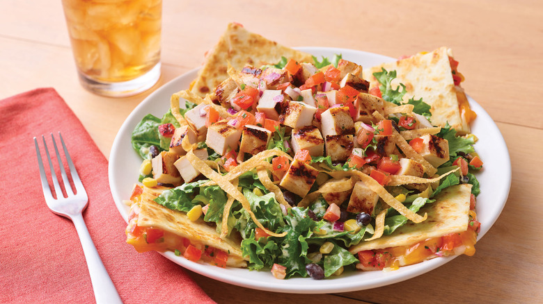 white plate of quesadilla chicken salad on a wooden table near a glass of iced tea with a fork on a red cloth napkin