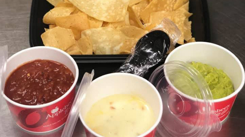 black plastic container of nacho chips with a plastic cup of white queso, a cup of salsa, and a cup of guacamole