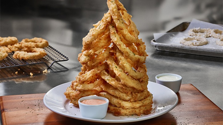 A tower of Homemade Onion Rings with dipping sauces is shown on a plate with other onion rings on a grate and sheet pan in the background.