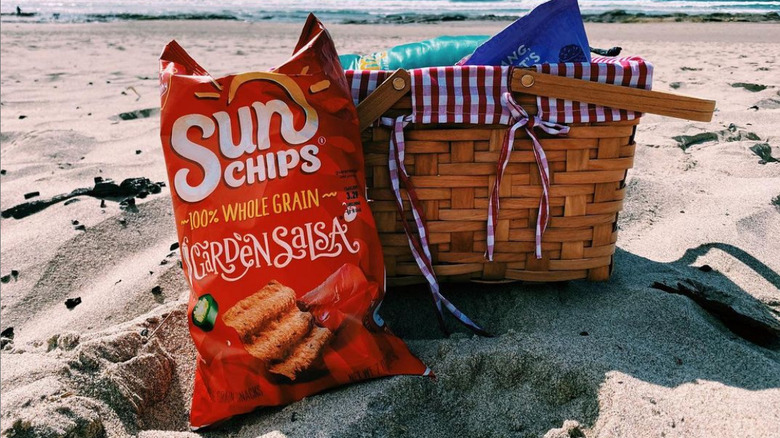 A bag of garden salsa sun chips sitting in the sand at the beach next to a picnic basket