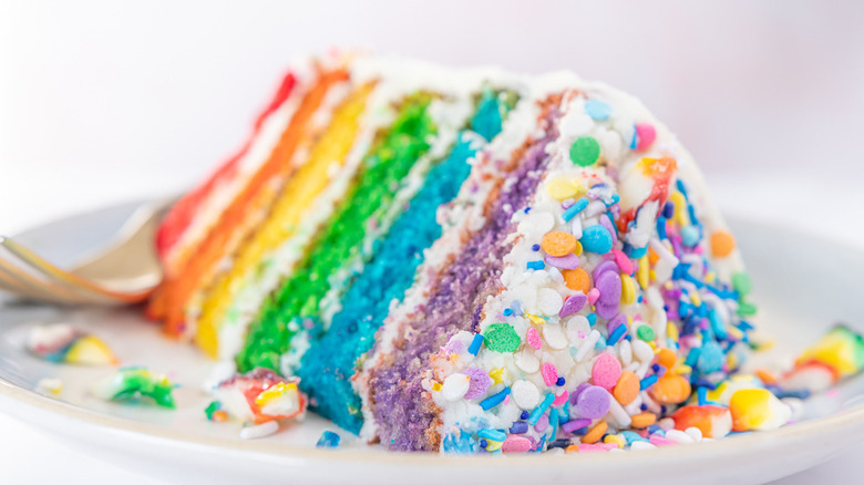 A slice of rainbow cake made with six layers of colored cake, white frosting, and rainbow sprinkles