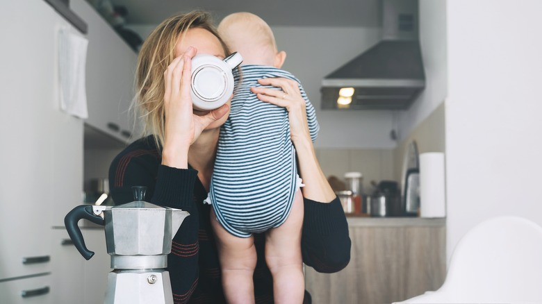 Tired mother holding baby in one hand and drinking mug of coffee in the other hand