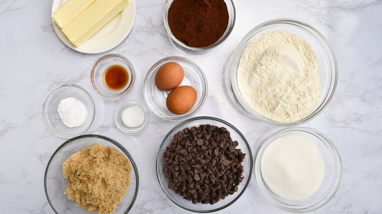 Glass bowls filled with chocolate chips, cocoa, flour, sugar, vanilla, baking soda, salt and eggs next to a plate with butter