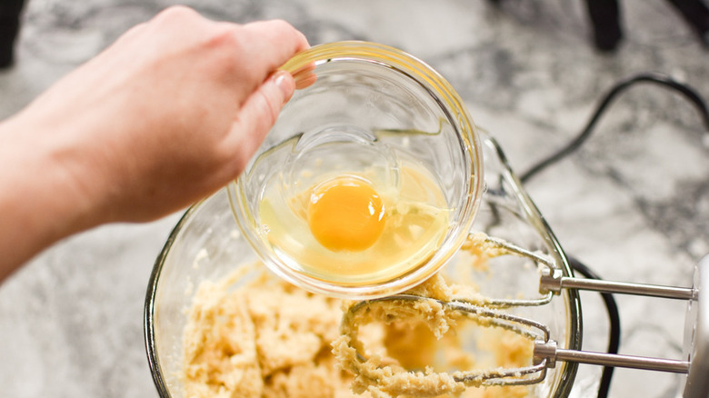 Close up of a hand holding an egg in a glass bowl over a mixer's bowl