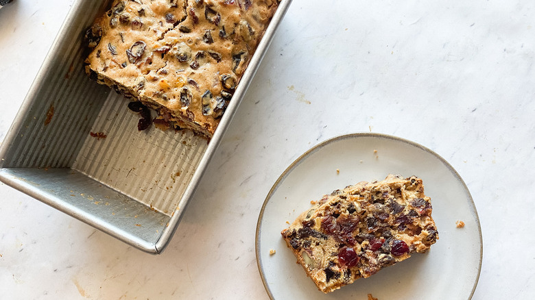 a slice of fruit cake on a plate with the loaf in a metal pan